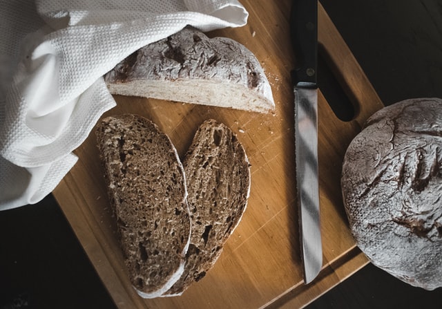 Auf einem Holzbrett liegt ein aufgeschnittenes Brot mit einem Messer, einem Tuch und zwei abgeschnittenen Scheiben
