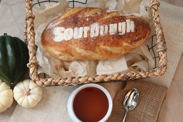 In einem Brotkorb liegt ein helles Sauerteigbrot, auf dem mit Mehl "Sourdough" geschrieben steht. Daneben liegt etwas Gemüse, ein Löffel und eine Tasse mit Tee